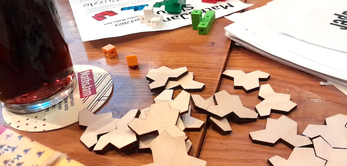 Photo of some objects on the table in a pub, including card games, aperiodic tiles, puzzle sheets and a pint of coke.